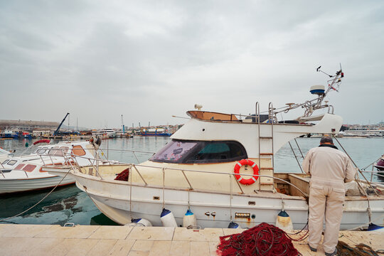 A man stands near a docked fishing boat with nets, in a harbor setting.