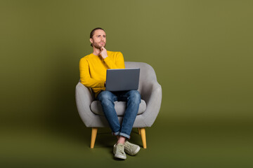 Young man in a casual outfit sitting on a chair holding a laptop, deep in thought with a contemplative expression