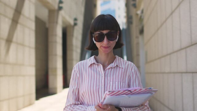 A businesswoman wearing headphones answers a call and takes notes in a notebook. A portrait of a confident, elegant businesswoman wearing headphones talking on the street. 