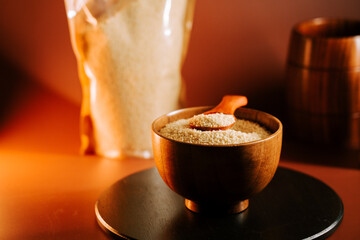 Uncooked couscous in a wooden bowl with a spoon on a dark surface beside a bag
