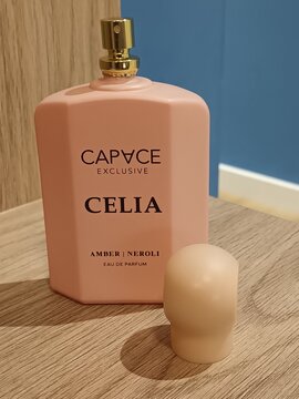Pink perfume bottle labeled 'CELIA' beside its cap on a wooden surface with a blue wall background