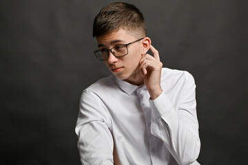 Young man wearing glasses and white shirt posing in thoughtful mood, turning head slightly, hand touching ear, expressing introspective feeling. Mood thoughtful, posture thoughtful.