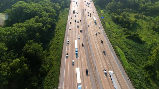 Highway road with the fast-moving vehicles, trucks showing U.S. transportation infrastructure under the bright sky in green suburbs. Drone view.