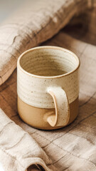 A rustic ceramic mug with a speckled beige glaze and sturdy handle rests on soft linen fabric, evoking warmth, coziness, and relaxed mornings.