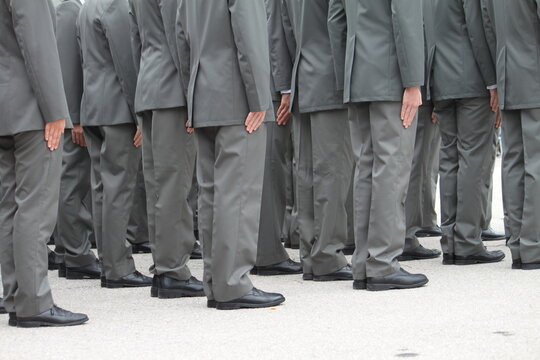 A group of people in gray uniforms stand in formation with their hands on their trouser seams. No heads or faces are visible. They are all identical, standing at attention. Rear view from behind.