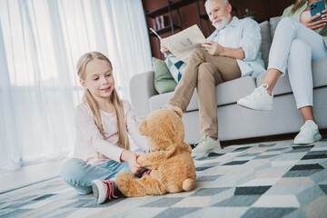 Playful moment at home as a young girl shares a teddy bear while parents relax on the sofa creating a warm family scene in a cozy living room