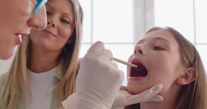 ENT doctor in gloves examining throat of child on otolaryngology appointment, mother standing nearby during examination of girl in clinic. Kid sitting with open mouth with trust in pediatrician