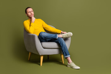 Young man wearing casual yellow shirt sitting thoughtfully on a modern armchair in front of a solid green background
