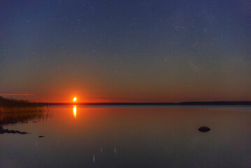 The moon sets behind the horizon against the backdrop of a lake.