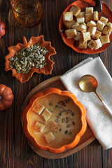 Creamy pumpkin soup with croutons and seeds, served in a pumpkin-shaped bowl on dark wooden table. Top view
