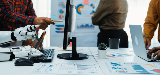 A dynamic workspace showcases a human collaborating with a robot, both engaged with computers, displaying vibrant data charts. This modern office symbolizes innovation and teamwork.Noogenesis