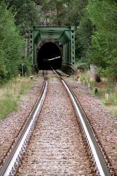 Train tracks leading into a dark tunnel entrance. A perspective view of railway tracks approaching a tunnel portal framed by dense forest foliage on a bright day