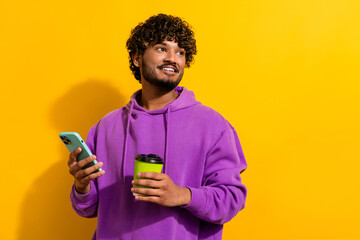 Young stylish man holding coffee cup and phone on a vibrant yellow backdrop, displaying a trendy and cheerful lifestyle