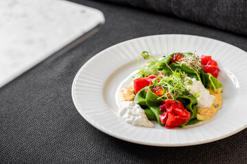 Gourmet dish with green crepes, microgreens, red vegetables, and creamy sauces on a white plate. Fine dining presentation on dark textile background. Horizontal, close-up.