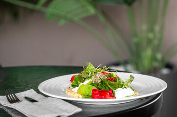 Fresh gourmet salad with microgreens, roasted peppers, and creamy dressing on a white plate, styled on a dark green table with tropical leaves and napkin for elegant presentation.