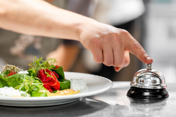 Close-up of a hand pressing a service bell near a gourmet plated dish in a restaurant or hotel setting, highlighting hospitality, fine dining, and professional service.