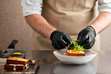 Chef in apron and gloves garnishing toasted sandwich with fresh arugula in professional kitchen. Croque monsieur on plate, egg in muffin tin, stainless steel counter.