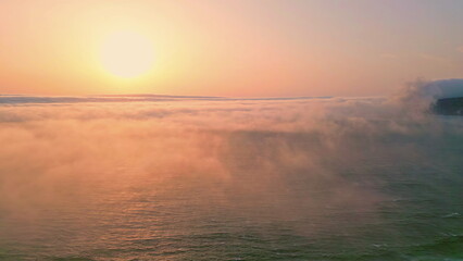 Amazing ocean sunset illuminating white fog covering calm water aerial view. 