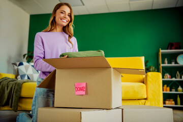 Young woman in living room donating clothes in a box, supporting charity, joyful expression, positive atmosphere, modern interior