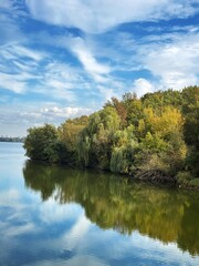 Calm river with reflections of clouds and trees under a blue sky.