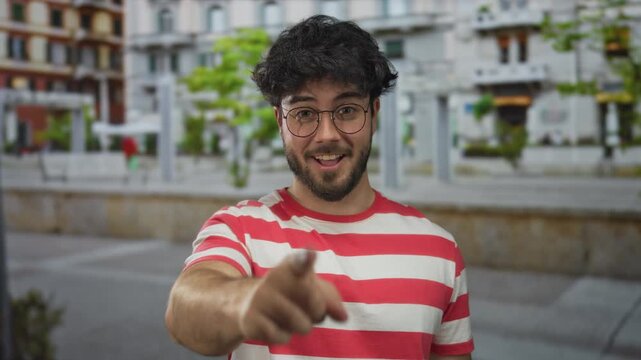 Young man with beard smiling outdoors on a street wearing glasses in a red striped shirt pointing happily on a bright day embracing fun and confidence with vibrant ambiance.