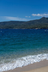 Scenic coastal view with turquoise sea waves washing sandy shore, lush green mountains in the background under clear blue sky.