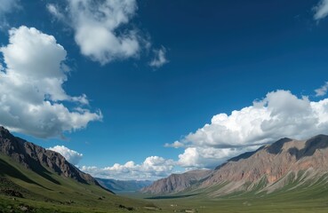 Vast green valley with rolling hills and mountains under a bright blue sky filled with fluffy white clouds. Sunlight casts shadows on the slopes, revealing rugged terrain. Remote, natural landscape.