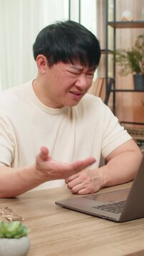 Asian man sitting at home table rereads bad email on laptop, angrily tapping keyboard, brow deeply furrowed. Chinese guy shows tense frustration, shoulders rigid, disappointment and stress visible.