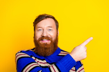 Fototapeta premium Smiling red-haired man with beard pointing right against yellow background wearing striped sweater.