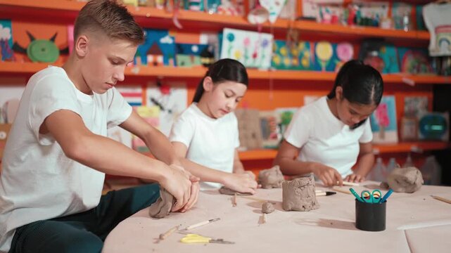 Caucasian smart teenager modeling clay while craving glass of clay at art lesson. Group of multicultural happy student working at dough in pottery workshop. Creative activity concept. Edification