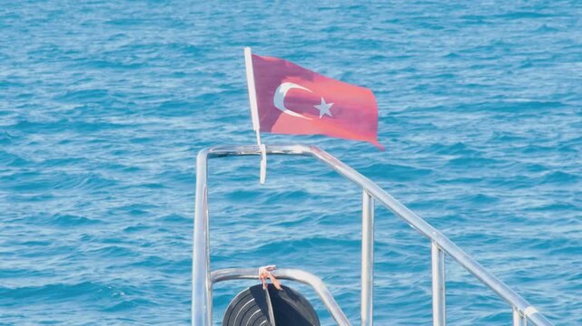 A Turkish flag (Ay Yıldız) proudly waving in the breeze on the railing of a boat or yacht. The background features the stunning, turquoise blue of the Mediterranean Sea. 