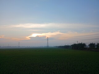 A serene landscape featuring a vast green field under a soft, gradient sky at dawn or dusk. The light is gentle, creating a peaceful and hazy atmosphere over the rural scenery.