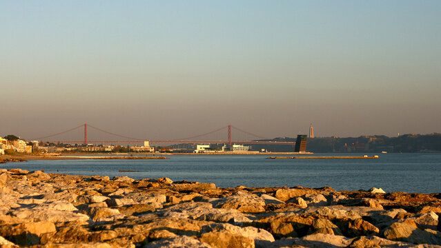 Golden Sunset Over Rocky Shore With Calm Water, Red Bridge, And Distant City Silhouette Beyond