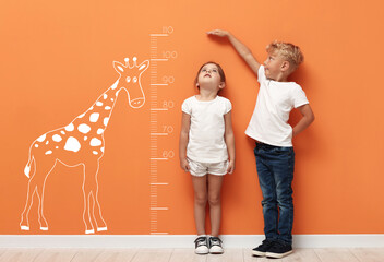 Cute little children measuring their heights near orange wall with scale and drawing of giraffe