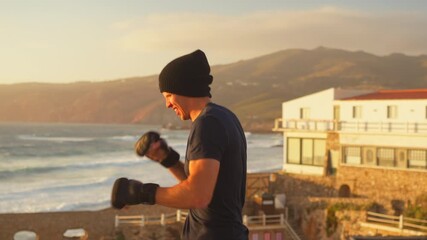 Fit young man practicing air boxing near seascape with mountains in the background at sunset, performing intense interval training in nature with focus and determination