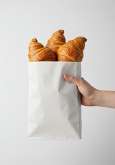 Hand holding a white paper takeaway bag filled with three golden-brown croissants on a clean background