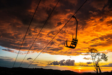 An inactive cable car frames a spectacular sunset, where hues of orange and purple dance across the sky. The wet silhouette of the cable car seat, along with the lone tree in the foreground
