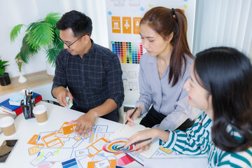 Fototapeta premium Three people are sitting at a table, working on a project. One of them is holding a piece of paper with a design on it. Scene is collaborative and focused 
