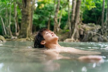 Fototapeta premium Young Asian man travel forest mountain hiking and camping on summer holiday vacation. Generation z guy enjoy active lifestyle relaxing in waterfall lagoon and looking beautiful nature in sunny day.
