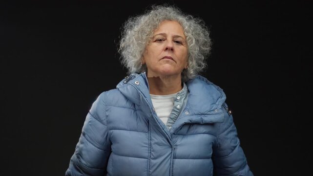 Woman with grey hair in blue coat gesturing on black background with a serious expression, capturing an isolated, elderly presence in a dramatic setting.