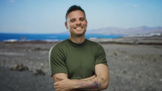 Young man with tattoos smiling confidently on a sunny beach, embracing a relaxed vibe with the ocean and clear sky in the background, showcasing a serene seaside atmosphere.