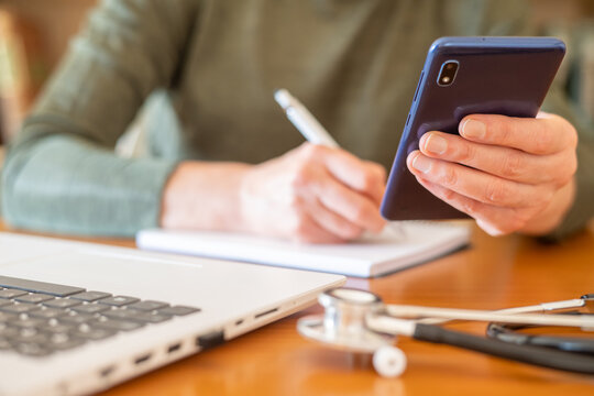 Medical professional studying healthcare information, taking notes while checking data on a smartphone, laptop and stethoscope on desk indicating remote learning or telehealth research