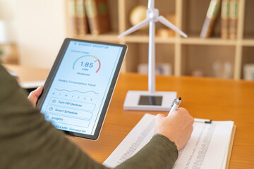 Person analyzing renewable energy consumption on a tablet, managing smart home devices and documents for sustainable living, with a desktop wind turbine model nearby