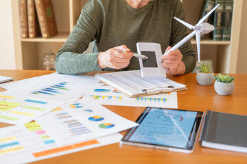 Woman hands holding a small solar panel and wind turbine model, analyzing charts and graphs on a desk with a tablet displaying energy data, symbolizing sustainable business planning