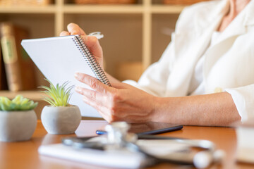 Doctor in white coat writing notes in spiral notebook at clinic desk with stethoscope, documenting patient consultation, diagnosis and treatment during primary care visit