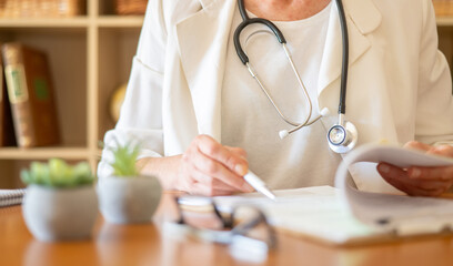 Healthcare professional in white coat with stethoscope reviewing patient charts and writing prescriptions at clinic desk, focused on paperwork and patient care