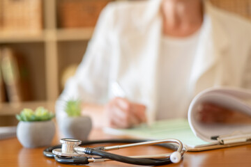 Stethoscope placed on a wooden desk while a blurred doctor in a white coat sits in the background, focusing on writing notes and reviewing documents on a clipboard