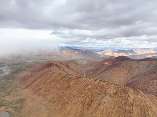 Aerial view over the textured, eroded ridges of high-altitude mountains fading into a hazy, wide valley under an overcast sky