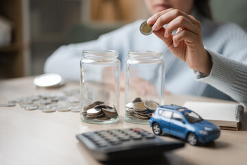 Hand placing a coin into glass jars on a desk, with calculator and toy car, illustrates an emergency fund and personal savings, highlighting safety cushion, risk management and budget planning.