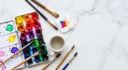 A watercolor paint set with vibrant hues several brushes a cup of clear water and a paint-stained tissue lie on a white marble surface